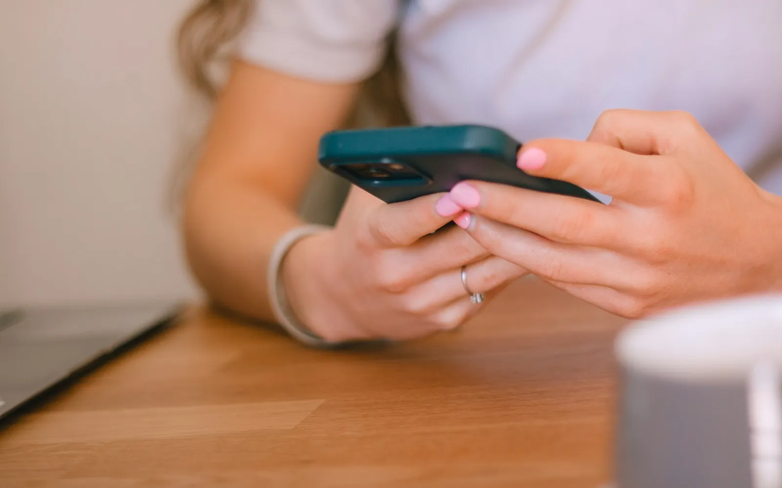 Hands holding a phone over a wooden table, with a laptop and a mug nearby